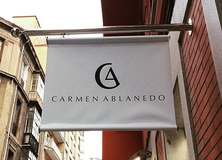 Vista del cartel exterior, perpendicular a la fachada del comercio, dónde vemos el logotipo y el nombre del comercio «Carmen Ablaneo»