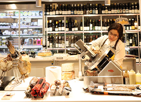 Una dependiente corta un producto en el mostrador donde podemos ver embutidos y jamones dispuestos para su corte a cuchillo. Al fondo se ve una estantería con vino y un expositor refrigerado con diversos productos