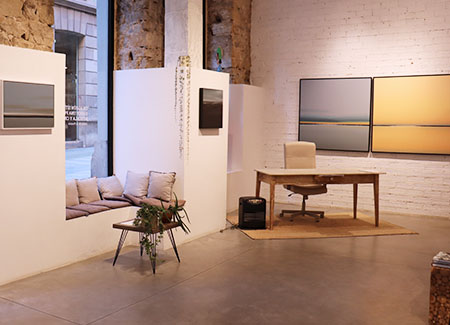 Vista del interior de la galería, dónde podemos ver una mesa de oficina en una esquina, un ventanal con cojines para sentarse frente a una mesa con una planta y cuatro cuadros en las paredes, dos pequeños a ambos lados del ventanal y dos grandes en la otra pared