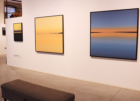 Vista del interior de la galería dónde podemos ver tres cuadros, alineados en una pared, dos grandes y uno pequeño. En primer plano se vislumbra un banco para sentarse