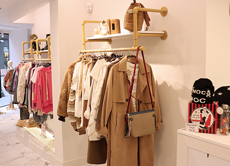 Vista del interior de la tienda, dónde podemos ver dos percheros separados por un saliente de la pared, con estante, en los que tenemos diversas prendas de ropa en perchas, así como botas y bolsos en los estantes superiores