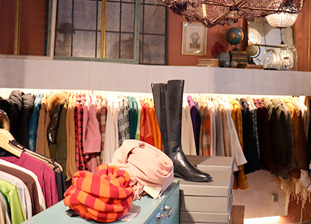 Vista del interior de la tienda, donde podemos ver un gran perchero de pared con múltiples prendas de ropa, en primer plano vemos unas bufandas y unas botas altas sobre expositores