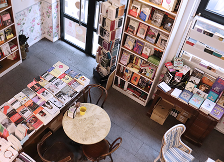 Vista cenital de la planta baja de la librería, dónde podemos ver la puerta de entrada, estanterías con libros, dos expositores con libros, una mesa con tres sillas de madera y una silla acolchada