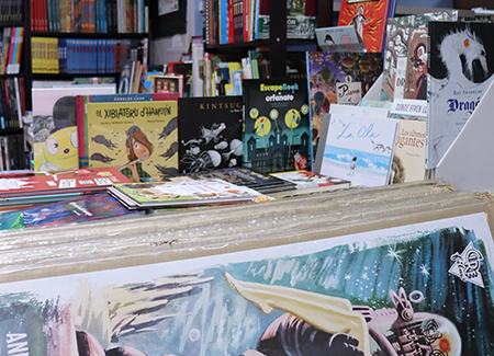 Vista del interior de la librería dónde podemos ver un expositor con cómics, al fondo un estantería con libros y en primer plano vemos parcialmente una colección de carteles de películas protegidos por cartón y plástico que están tumbados