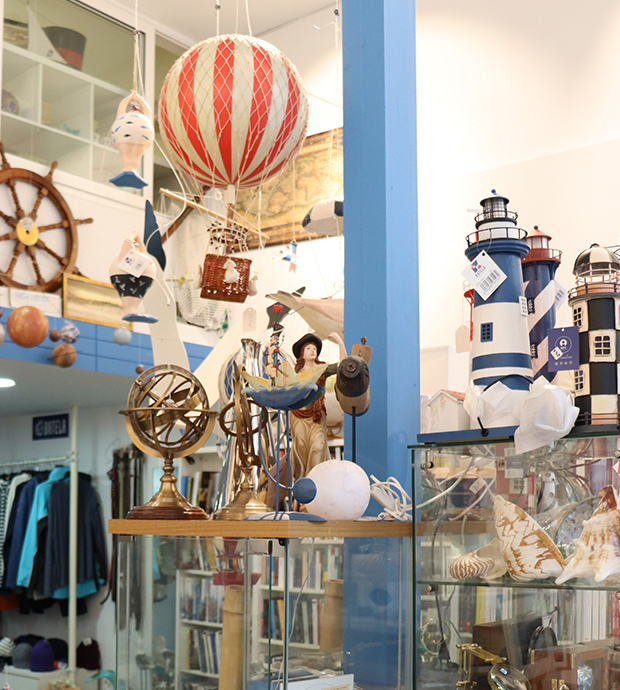 Vista del interior de la librería, dónde pdemos ver unas vitrinas con artículos de decoración (faros, globos aerostáticos, conchas...), al fondo se ve un perchero de tipo burro con ropa