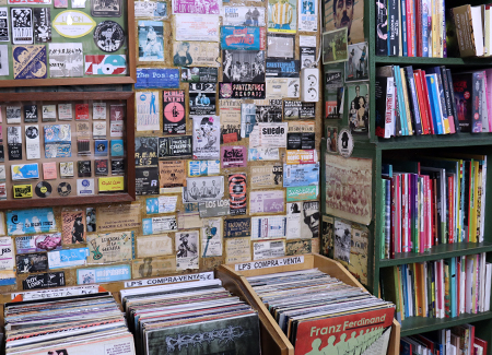 Vista del interior de la librería, dónde vemos un expositor de discos de vinilo, en la pared de detrás podemos ver fotos, postales, carteles y dos marcos con recuerdos. A la derecha de la foto se ve una estantería con libros.