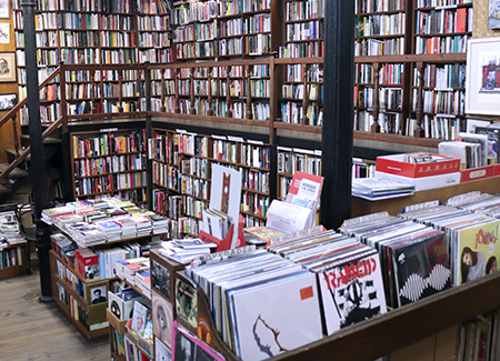 Vista del interior de la librería, dónde vemos una esquina del comercio cubierta por estanterías con dos niveles de altura y una escalera que lleva al nivel superior. Frente a la estantería hay un expositor con más libros y perpendicular a ella otro expositor con más libros y con discos de vinilo.