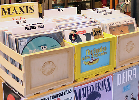 Primer plano de un expositor con vinilos de distintos artistas y estilos en cajas de madera. Del expositor cuelgan posters de artistas