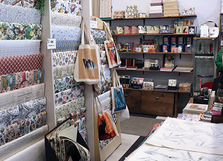 Vista del interior de la papelería, dónde podemos ver a la izquierda un expositor vertical de papel de regalo, en el que también tenemos bolsas de tela, en primer plano vemos láminas de dibujo y al fondo una estantería con material de regalo