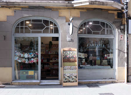 Vista del exterior de la papelería, dónde podemos ver la entrada y los dos escaparates, uno adyacente a la puerta, junto a un expositor