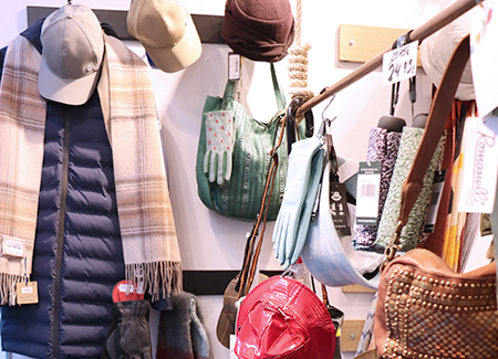 Vista del interior de la tienda, dónde se puede ver en dos percheros, uno de pie y otro de pared, diversos complementos: bolsos, guantes, gorros, bufandas, paraguas...