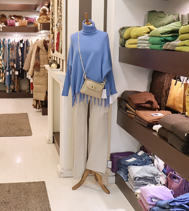 Vista del interior del comercio, dónde podemos ver a la derecha unas baldas con ropa de doblada de diferentes colores. A su lado un maniquí que luce una falda veis, un poncho azul y un bolso cruzado. Al fondo se ven percheros con más ropa