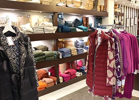 Vista del interior de la tienda dónde vemos en primer término dos percheros de tipo burro, con ropa de diferentes colores y estilos. Al fondo un estantería con ropa doblada