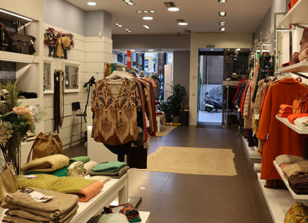 Vista del interior de la tienda, dónde se puede ver un pasillo con estanterías y percheros de pie a un lado, con un expositor y otro perchero de pie al otro lado. Al fondo se ve la entrada a la tienda