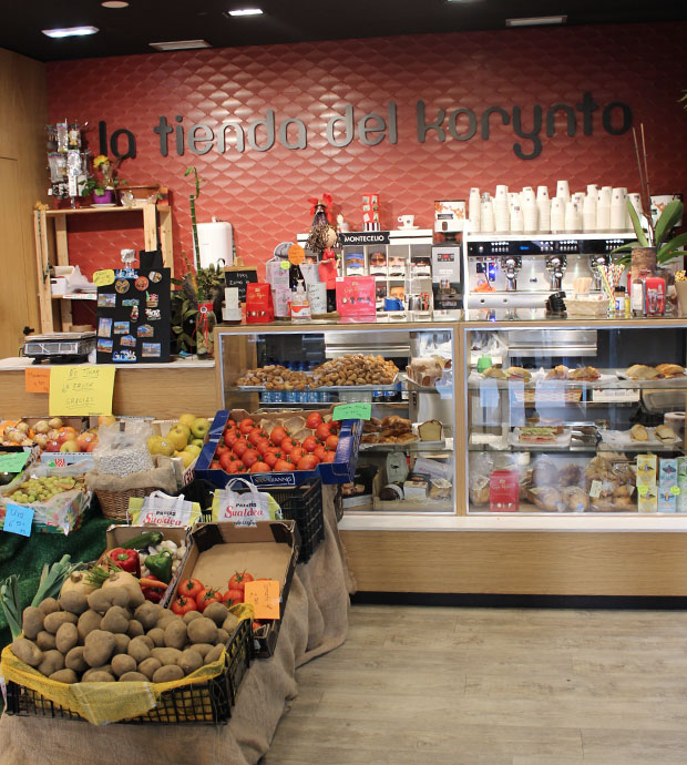 Vista del interior de la tienda, dónde podemos ver el mostrador expositor con diversos productos, tanto dentro como sobre el, a su lado hay un mostrador con adornos y recuerdos. En primer plano vemos un expositor con frutas y verduras, así como un saco con faba asturiana. Al fondo vemos una cafetera profesional, cubierta por tazas y estanterías con más productos